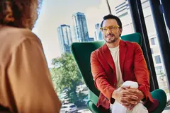 Male with glass and red blazer sitting in a green chair looking at a person