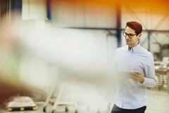 Male worker in a tech environment. Holding papers. Wearing glasses. Factory/logistics. Solid white shirt. Primary color cream. Secondary color yellow/orange.