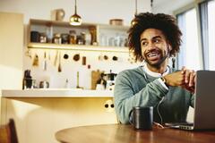 Smiling man sitting at his dining table with a drink and his laptop. Looking away. Kitchen in the background.
