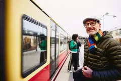 male standing next to a train. Two people getting out of a train in the background.
