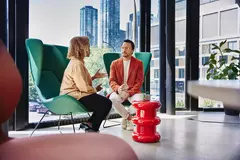 Male with glass and red blazer sitting in a green chair talking to a female sitting in a green chair