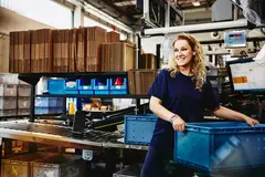Smiling female standing next to conveyer belt holding a crate
