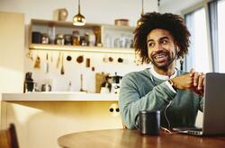 Smiling man sitting at his dining table with a drink and his laptop. Looking away. Kitchen in the background.