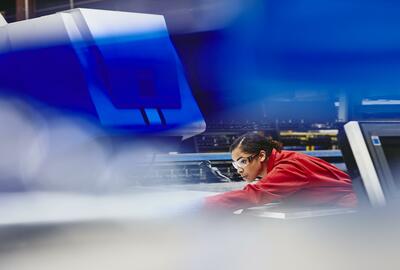 Female blue-collar worker aligning something. African-american woman.  Wearing safety goggles and an overall. Focused. Ponytail. Overall. Primary color blue. Secondary color red.