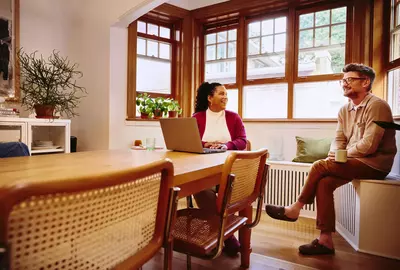 Female sitting at a table with laptop talking to male with coffee mug at home
