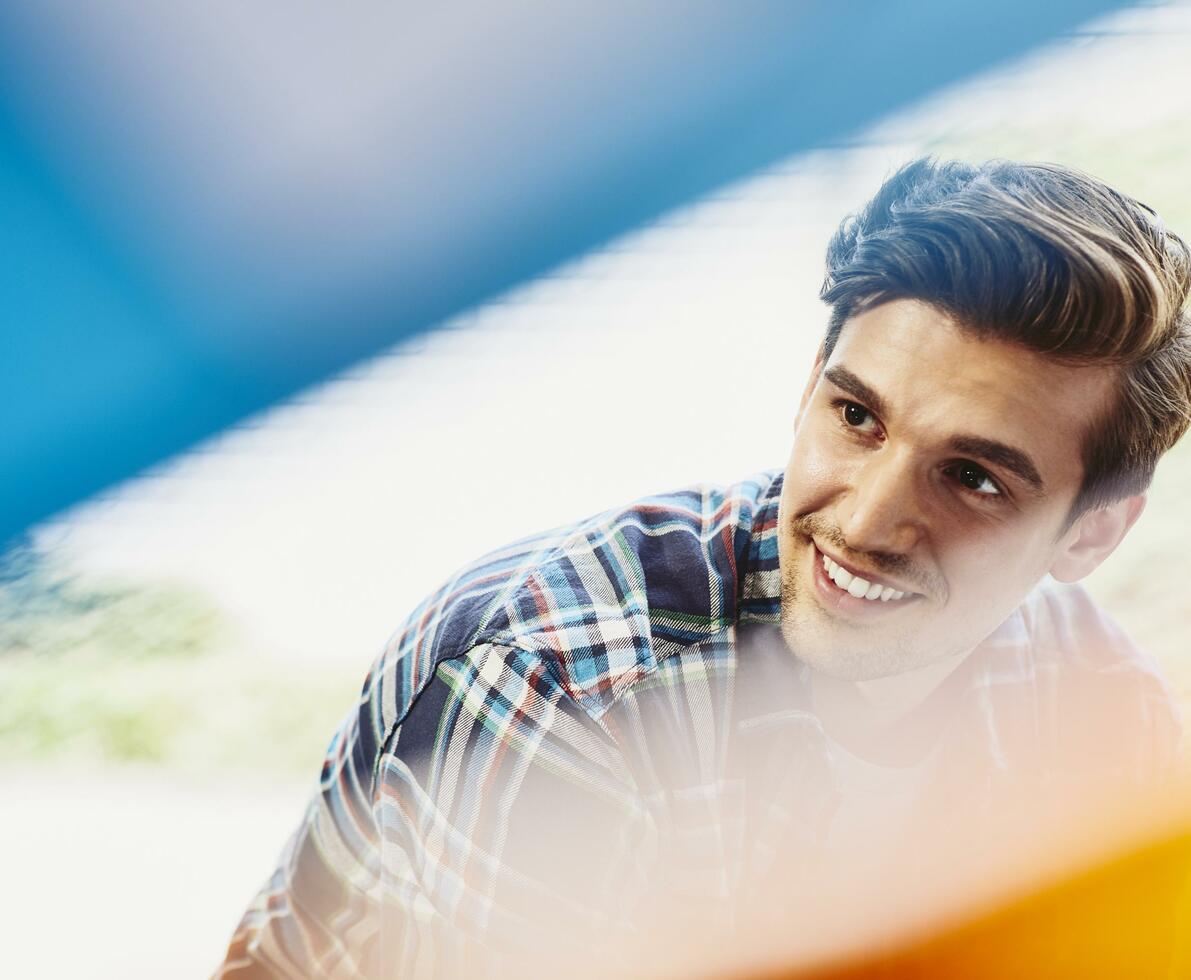 Male employee smiling and looking to his right. Groomed beard and mustache. Wearing a checkered shirt. Primary color blue. Secondary colors cream and yellow.