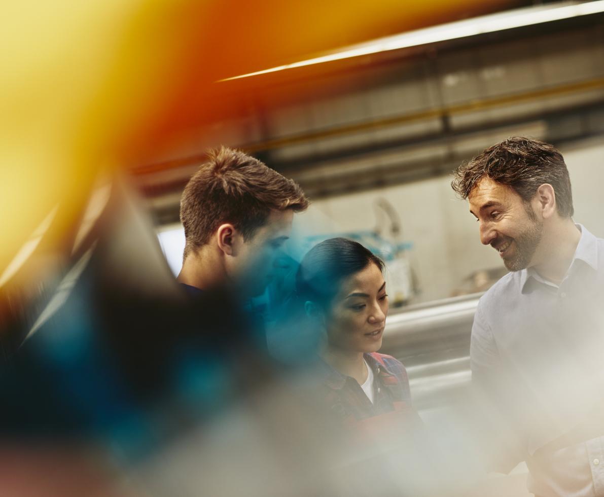 People in a tech environment discussing. Two males and one female. Caucasian (white) men and Asian woman. Primary color yellow. Secondary color blue.