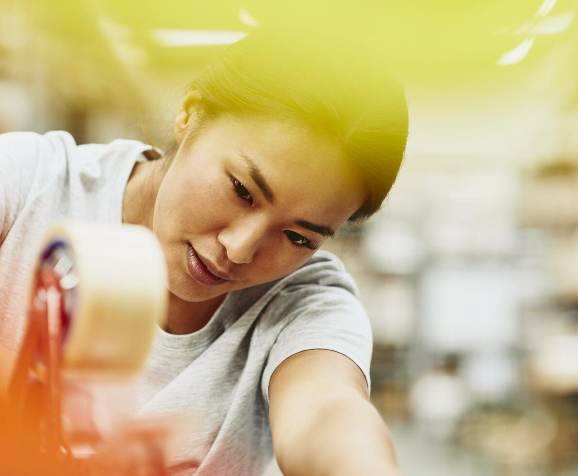 Female blue-collar worker focused on taping a box. Logistics environment. Hair up. Asian woman. Primary color yellow. Secondary color  orange.