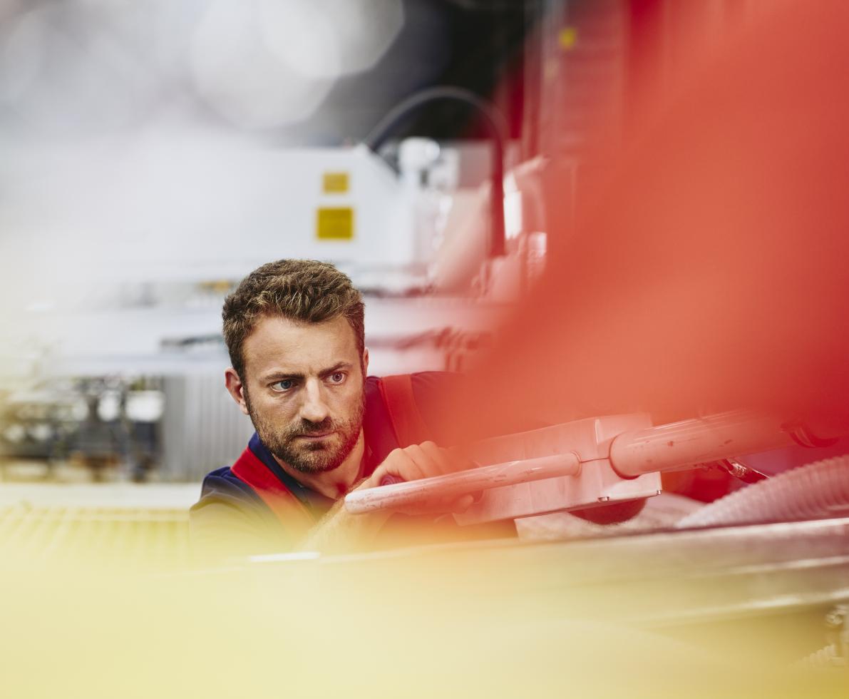 Blue-collar worker aligning something. Very focused. Caucasian man. Groomed beard. Primary color red. Secondary color cream/yellow.